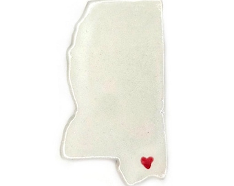 May include: A white, translucent Mississippi state-shaped ornament with a red heart marking a location. The ornament has a slightly textured surface and a clear, raised border. The heart is a solid, vibrant red.