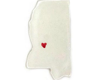 May include: A white, irregularly shaped object in the form of the state of Mississippi, with a small red heart marking a location within the state. The object has a slightly translucent appearance and a smooth surface.