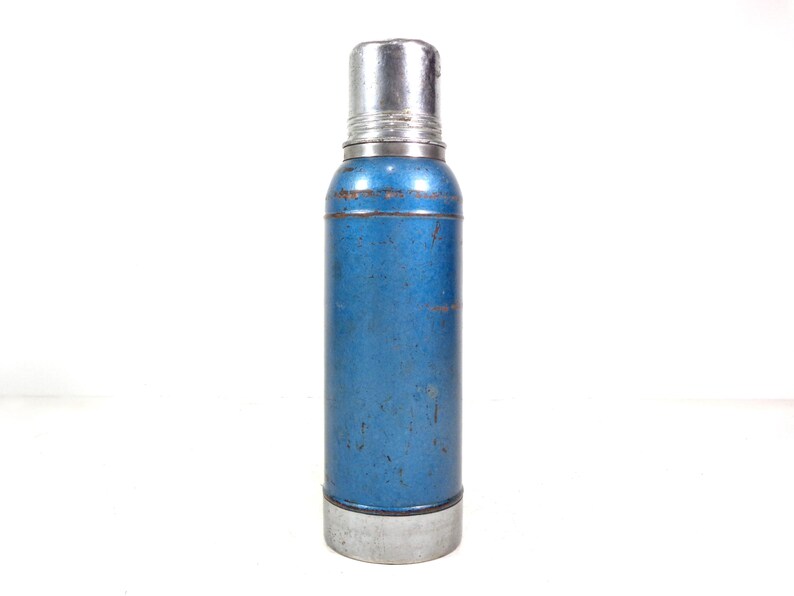 Pode incluir: Uma garrafa t&eacute;rmica azul e prateada vintage. O corpo cil&iacute;ndrico &eacute; azul desbotado com a parte superior e inferior prateadas. A garrafa t&eacute;rmica tem uma tampa de rosca e parece ser feita de metal. O design geral sugere um estilo cl&aacute;ssico e retro.