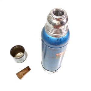Pode incluir: Uma garrafa t&eacute;rmica azul e prateada vintage com tampa e copo prateados. Uma rolha de corti&ccedil;a castanha est&aacute; ao lado do copo. A garrafa t&eacute;rmica tem um corpo azul com algum desgaste. O copo &eacute; prateado e tem uma borda enrolada.