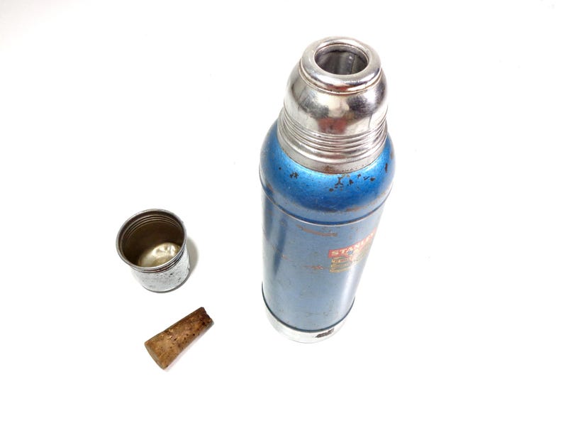 Pode incluir: Uma garrafa t&eacute;rmica azul e prateada vintage com tampa e copo prateados. Uma rolha de corti&ccedil;a castanha est&aacute; ao lado do copo. A garrafa t&eacute;rmica tem um corpo azul com algum desgaste. O copo &eacute; prateado e tem uma borda enrolada.
