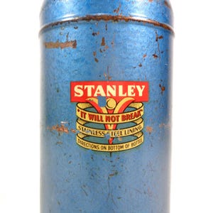 Pode incluir: Termo Stanley azul vintage com acabamento desgastado. O r&oacute;tulo apresenta o nome "STANLEY" em vermelho e dourado, com o texto "IT WILL NOT BREAK" e "STAINLESS STEEL LINING". O termo tem formato cil&iacute;ndrico e um design cl&aacute;ssico.