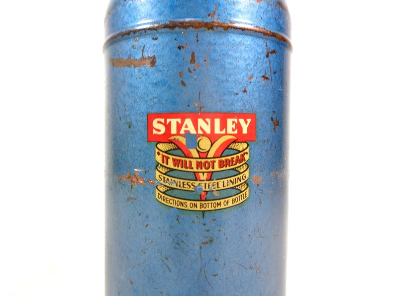 Pode incluir: Termo Stanley azul vintage com acabamento desgastado. O r&oacute;tulo apresenta o nome "STANLEY" em vermelho e dourado, com o texto "IT WILL NOT BREAK" e "STAINLESS STEEL LINING". O termo tem formato cil&iacute;ndrico e um design cl&aacute;ssico.