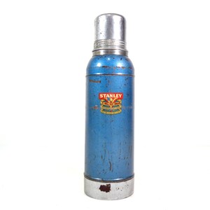 Pode incluir: Garrafa t&eacute;rmica Stanley vintage azul com tampa e base prateadas. A garrafa tem um r&oacute;tulo Stanley vermelho e dourado. O exterior mostra sinais de desgaste. Um recipiente isolado cl&aacute;ssico para bebidas.