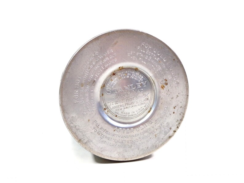 Pode incluir: Close-up da base de uma garrafa t&eacute;rmica Stanley Super Vac vintage. A base de metal prateado apresenta texto em relevo, incluindo "It will not break" e "Made in U.S.A.". As instru&ccedil;&otilde;es de uso tamb&eacute;m s&atilde;o vis&iacute;veis.
