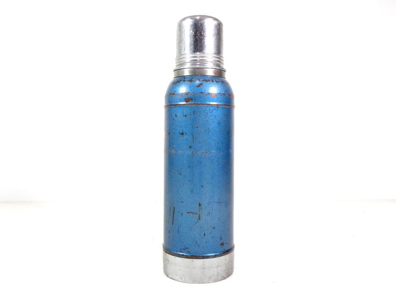 Pode incluir: Termo de metal azul vintage com tampa e base prateadas. A garrafa cil&iacute;ndrica mostra sinais de desgaste, com arranh&otilde;es vis&iacute;veis e uma cor ligeiramente desbotada. O termo foi projetado para manter l&iacute;quidos quentes ou frios.
