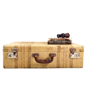 May include: A rectangular beige suitcase with vertical stripes, featuring a brown handle and two silver metal latches. Resting on top of the suitcase are two weathered, dark-colored vintage books and a pair of small, dark brown binoculars.