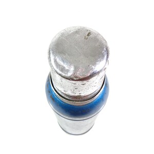Pode incluir: Um tamp&atilde;o de garrafa de metal vintage com uma parte superior prateada e uma faixa azul. O tamp&atilde;o tem uma forma cil&iacute;ndrica e parece ser feito de metal. A imagem &eacute; tirada de cima sobre um fundo branco.