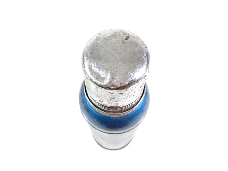 Pode incluir: Um tamp&atilde;o de garrafa de metal vintage com uma parte superior prateada e uma faixa azul. O tamp&atilde;o tem uma forma cil&iacute;ndrica e parece ser feito de metal. A imagem &eacute; tirada de cima sobre um fundo branco.