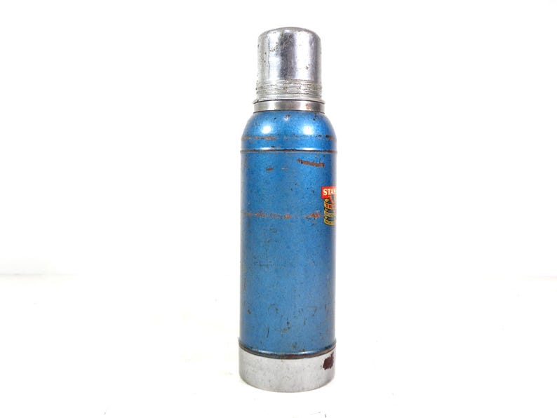 Pode incluir: Uma garrafa t&eacute;rmica Stanley azul vintage com tampa e base prateadas. A garrafa t&eacute;rmica tem formato cil&iacute;ndrico e um pequeno r&oacute;tulo com texto. O exterior mostra sinais de desgaste, indicando sua idade.