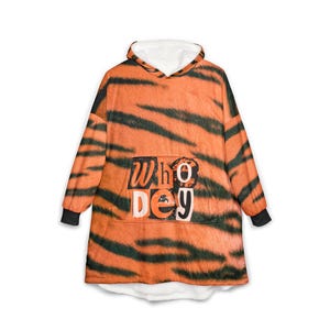 Pode incluir: Uma camisola com capuz oversized com um padrão de riscas de tigre em laranja e preto. A peça inclui um bolso frontal grande com o texto "Who Dey" impresso numa fonte arrojada e desgastada. Tem mangas compridas com punhos pretos.