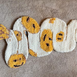 May include: A handmade rug in the shape of a snake, featuring a cream-colored base with yellow and black accents. The rug is made of a soft, textured material and is placed on a neutral-toned surface.