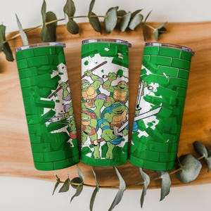 May include: Three green insulated tumblers featuring the Teenage Mutant Ninja Turtles. The tumblers have a green brick wall design and a graphic of the turtles in action. The tumblers are made of stainless steel and have a clear lid.