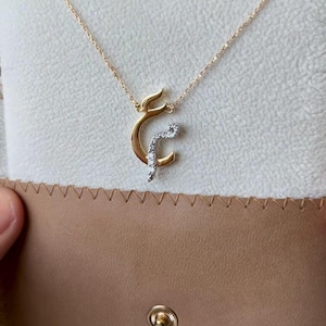 Puede incluir: Collar dorado con un colgante de caligrafía árabe y pequeñas piedras brillantes. El collar se muestra sobre un fondo neutro, dentro de un estuche de cuero marrón. La cadena es delicada y el colgante es el punto focal.