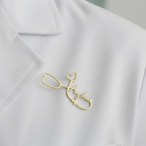 Puede incluir: Un broche de estetoscopio dorado con el nombre "Aysha" en escritura cursiva. El broche está sujeto a una prenda blanca, probablemente una bata de laboratorio o uniforme, y presenta un detalle en forma de corazón.