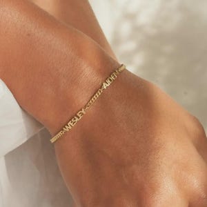 May include: A gold-colored chain bracelet with the names "WESLEY" and "AIDEN" engraved on it. The bracelet is worn on a wrist, and the chain has a delicate, textured design. The bracelet is a simple yet elegant piece of jewelry.