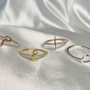 Könnte beinhalten: Vier Ringe mit arabischen Buchstabenmotiven. Die Ringe sind in Gold-, Roségold- und Silbertönen gehalten, jeder mit einem Band aus kleinen, klaren Steinen. Die Ringe werden auf einer weißen, strukturierten Oberfläche präsentiert.