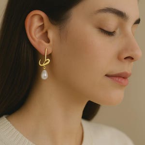 May include: Gold-toned earrings with a unique design, featuring a stylized script element and a dangling pearl. The earrings are worn by a person with dark hair and a cream-colored sweater. The background is a neutral beige.