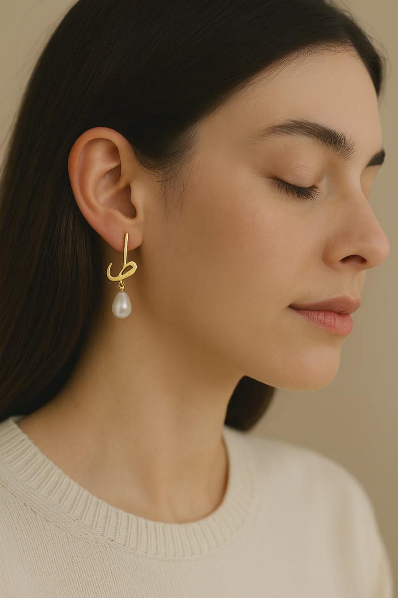 May include: Gold-toned earrings with a unique design, featuring a stylized script element and a dangling pearl. The earrings are worn by a person with dark hair and a cream-colored sweater. The background is a neutral beige.