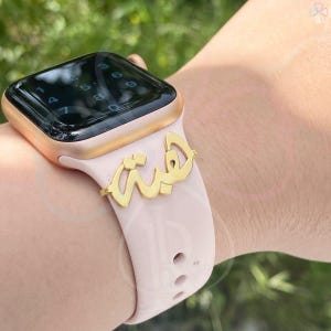 May include: A gold-coloured charm with Arabic script is attached to a light pink Apple Watch strap. The watch has a gold case and a black screen displaying numbers. The strap is secured on a wrist.