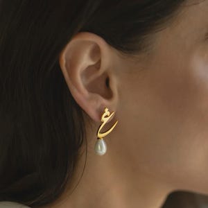 May include: A gold-colored earring in the shape of Arabic script is shown. The earring has a teardrop-shaped pearl pendant. The earring is worn on a person's ear, with the person's shoulder and neck visible in the background.