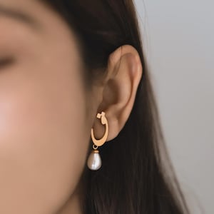 May include: Gold-toned hoop earring with a teardrop pearl pendant. The earring is worn on the ear of a person with dark hair. The pearl is white and has a smooth surface. The earring has a unique design.