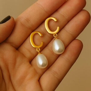 May include: Gold-toned earrings featuring a 'C' shape design with a dangling pear-shaped pearl. The earrings have a polished finish and a classic, elegant style. The pearls are white and have a smooth, lustrous surface.