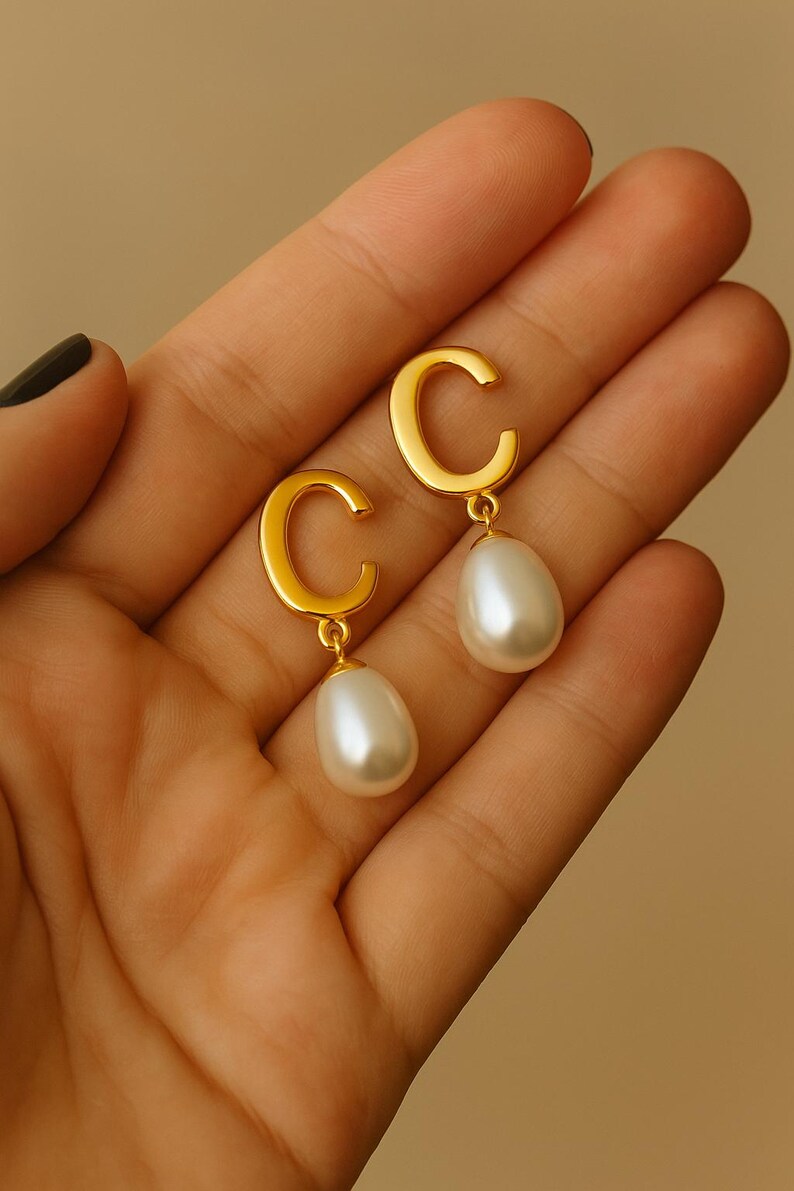 May include: Gold-toned earrings featuring a 'C' shape design with a dangling pear-shaped pearl. The earrings have a polished finish and a classic, elegant style. The pearls are white and have a smooth, lustrous surface.