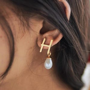 May include: Gold-colored letter "H" earrings with a pear-shaped, white faux pearl drop. The earrings are hanging from a person's ear, with the focus on the jewelry. The earrings are a simple, elegant design.