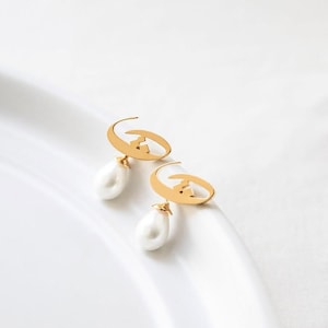 May include: A pair of gold-colored earrings with a crescent moon design and a dangling white teardrop-shaped pearl. The earrings are displayed on a white surface, showcasing their elegant and minimalist style.