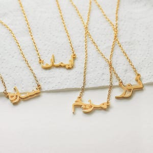 May include: Gold-colored name necklaces with Arabic script spelling out various names. The necklaces are displayed on a white surface. Each necklace features a delicate chain and a pendant with a unique name.
