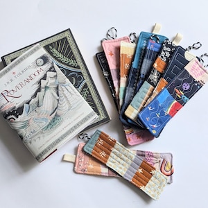 May include: A hardback book titled Roverandom by J.R.R. Tolkien next to a collection of rectangular fabric bookmarks. The bookmarks feature various patterns including space themes, rainbows, and geometric designs, each with a small fabric loop.