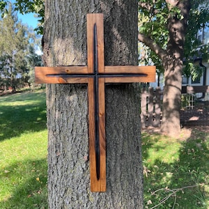 May include: A wooden cross with a dark, charred center and a natural wood finish. The cross is mounted on a tree trunk, showcasing the rustic texture of the wood and the tree bark.