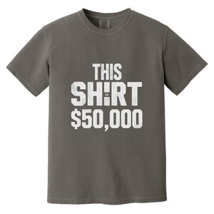 Puede incluir: Una camiseta gris con el texto "THIS SHIRT IS $50,000" en letras blancas desgastadas. La camiseta es de cuello redondo y parece estar hecha de un material suave y c&oacute;modo. La est&eacute;tica general es informal y humor&iacute;stica.