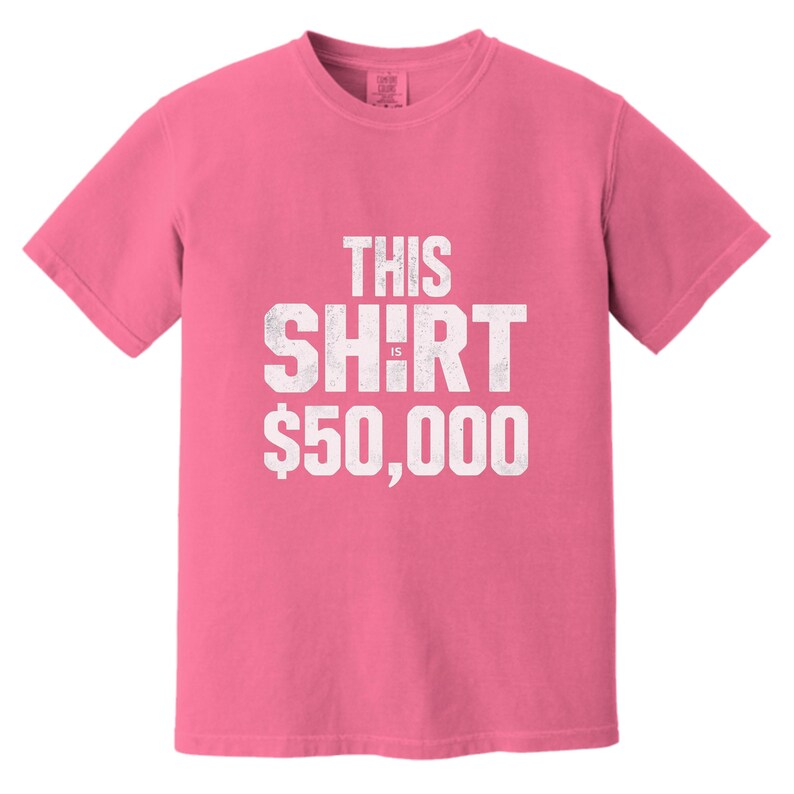 Puede incluir: Una camiseta rosa con el texto blanco "THIS SHIRT IS $50,000" en letras desgastadas. La camiseta tiene mangas cortas y cuello redondo. La tela parece ser una mezcla de algod&oacute;n suave.