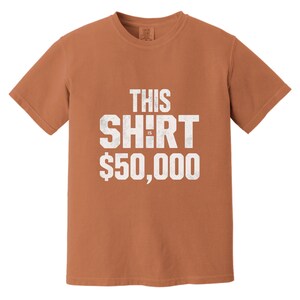 Puede incluir: Una camiseta de color &oacute;xido con el texto "THIS SHIRT IS $50,000" en letras blancas desgastadas. La camiseta tiene cuello redondo y mangas cortas. El texto est&aacute; centrado en la parte delantera de la camiseta.