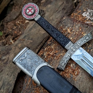 May include: A sword with a silver-toned crossguard featuring intricate scrollwork, a black grip with embossed knot patterns, and a circular pommel displaying a red cross. The accompanying black scabbard has a silver-toned metal fitting with woven relief designs.