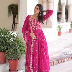 Puede incluir: Un vestido fucsia vibrante con intrincados bordados y adornos dorados. El vestido de manga larga presenta una silueta fluida, un corpiño detallado y una bufanda a juego. Adecuado para ocasiones especiales.