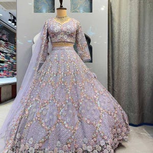 May include: A lilac embroidered formal gown with long sleeves and a matching veil. The gown features intricate floral patterns and embellishments. The bodice and skirt are adorned with detailed beadwork and sequins, creating a shimmering effect. The dress is displayed on a mannequin.