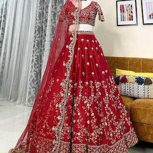 Op de afbeelding: Een levendige rode lehenga, een traditioneel Indiaas kledingstuk, met ingewikkelde gouden borduursels. De set bevat een bijpassende top en een doorschijnende dupatta. De rok heeft gedetailleerde bloempatronen en een decoratieve rand.