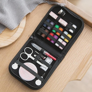 May include: A black sewing kit, open to display its contents. The kit includes spools of thread in various colors, a measuring tape, scissors, needles, pins, and a red pen. The kit is on a wooden surface.