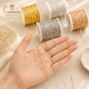 May include: Four spools of delicate satellite chain in gold, silver, and rose gold tones. Each spool holds a continuous length of thin metal chain featuring small, evenly spaced flat circular discs. The chains are displayed on white plastic spools.