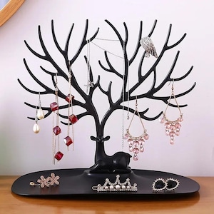 May include: Black metal jewelry organizer in the shape of a tree with a deer silhouette at the base. The organizer holds various earrings, necklaces, and hair accessories. The jewelry includes pearl, crystal, and rhinestone accents.