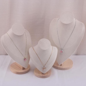 May include: Three necklace display busts with wooden bases. Each bust is covered in a light beige fabric and displays a different necklace. One necklace has a pink heart-shaped pendant, another has a star pendant, and the third has a round pink pendant.