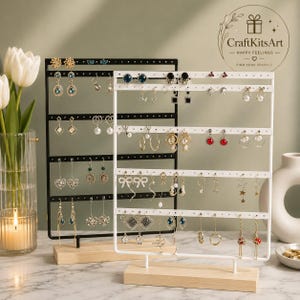 May include: Two rectangular metal earring display stands with light wood bases. One stand is black and the other is white, both featuring multiple horizontal rows with small holes for hanging various styles of earrings, including studs and dangles.