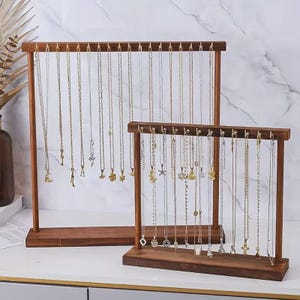 May include: Two wooden necklace display stands. The stands are made of dark brown wood and have a rectangular base and top with hooks to hang the necklaces. The necklaces are of varying lengths and feature different pendants. The stands are for displaying jewellery.