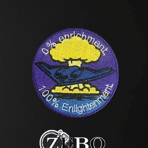 Könnte beinhalten: Gestickter Aufnäher mit lila Rand und einem zentralen Design mit einer gelben Atompilzwolke und einem stilisierten Flugzeug. Der Text lautet "0% enrichment" und "100% Enlightenment". Der Aufnäher enthält auch den Text "ZEBO".