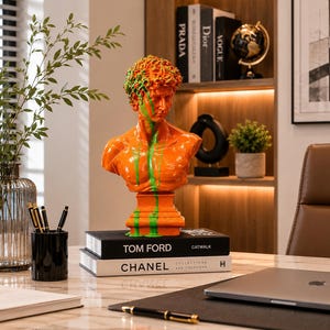 Pode incluir: Um busto laranja brilhante de uma figura masculina clássica com gotas de tinta verde vibrante sobre o cabelo, rosto e peito. A escultura está montada numa base quadrada e repousa sobre dois livros empilhados intitulados Tom Ford e Chanel.