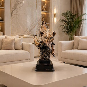 May include: A detailed sculpture of a leopard with a spotted coat, perched on a textured base. The sculpture is brown and gold, with a realistic depiction of the animal's face and fur. The sculpture is placed on a square, light-colored coffee table.