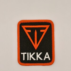 May include: A rectangular embroidered patch with a black background and a bright orange border. The patch features an orange triangular logo at the top and the word TIKKA embroidered in white capital letters centered at the bottom.